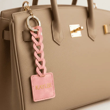 Pink chain keychain with Custom name tag attached to a beige handbag.