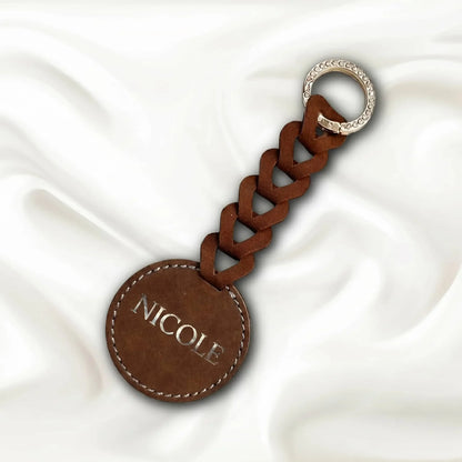 Brown leather keychain with braided design and 'NICOLE' engraving on a white fabric background